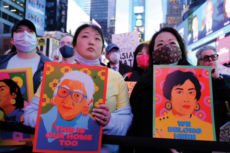 Two Asian women, one waring a face mask, at a rally holding signs with vibrant illustrations of Asian women. The signs say 