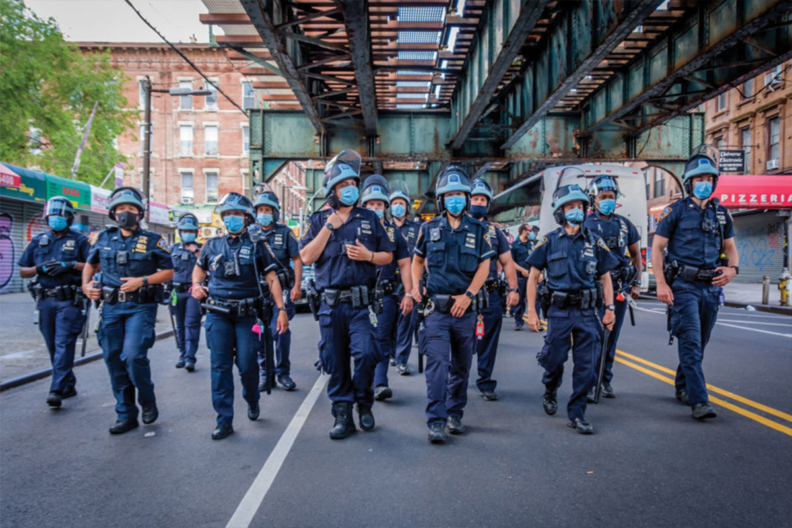 A large group of NYPD officers in riot gear and face masks walk down the street under an elevated train track